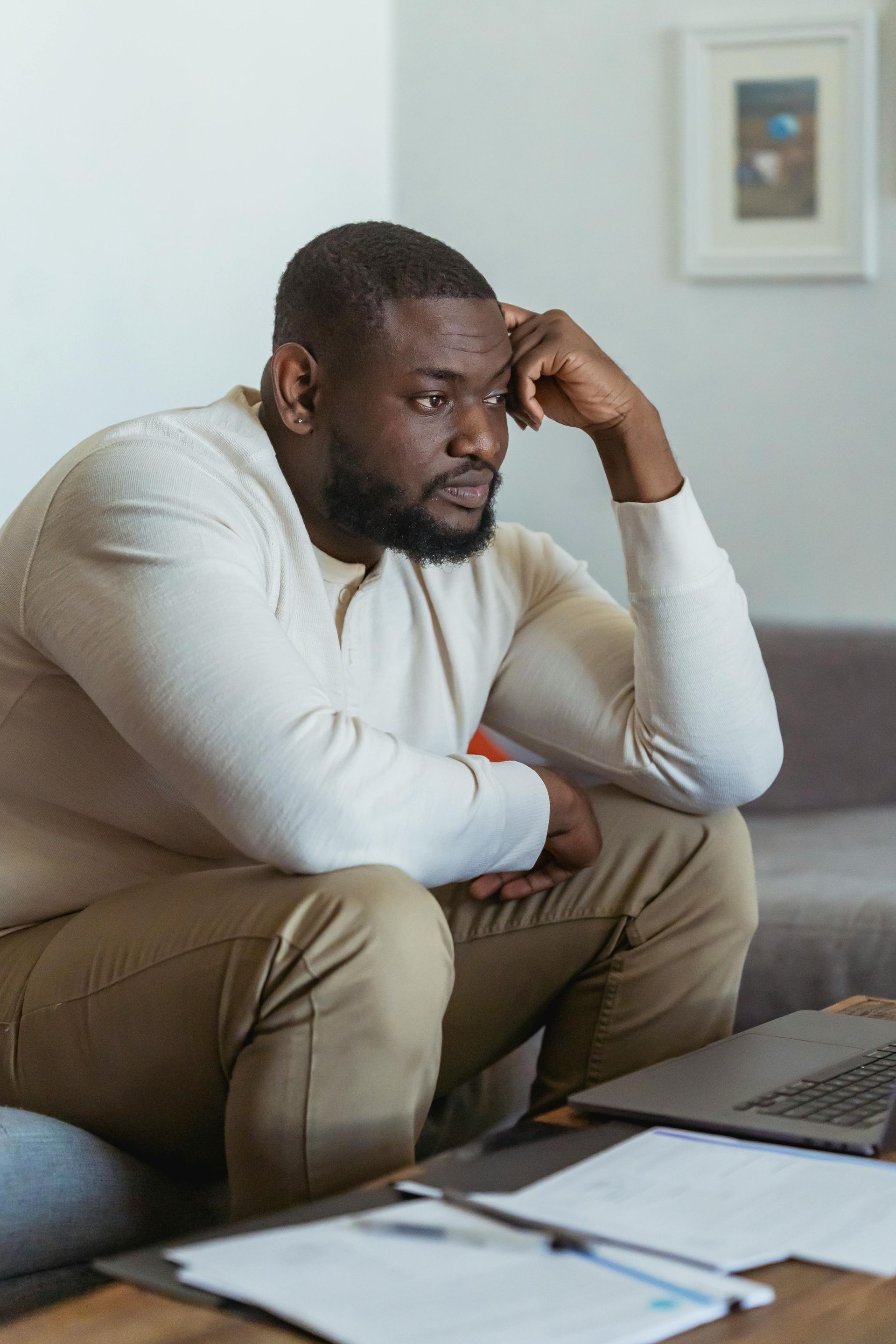 Man with beard, hand on head, looks down at papers and laptop, sitting on a sofa.