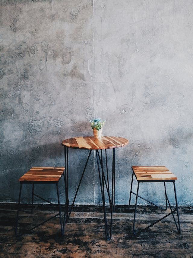 A wooden table and two stools in front of a concrete wall.