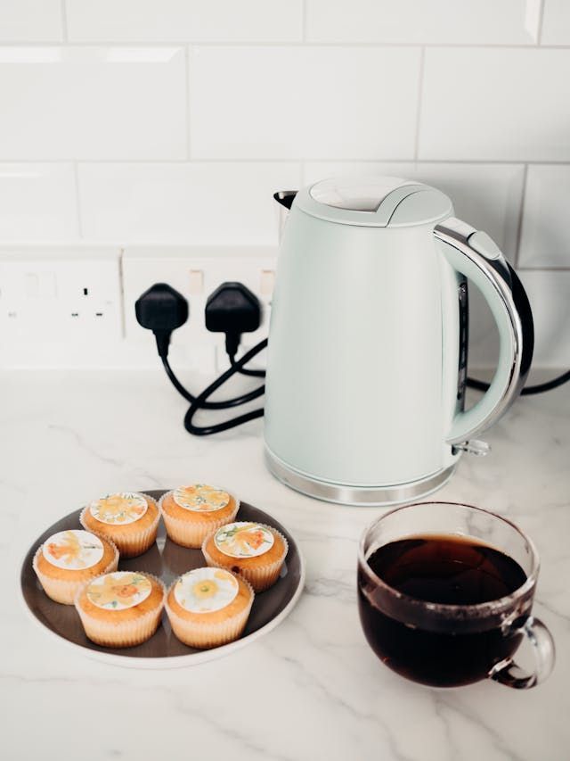A plate of pastries and a cup of coffee on a counter next to a kettle.