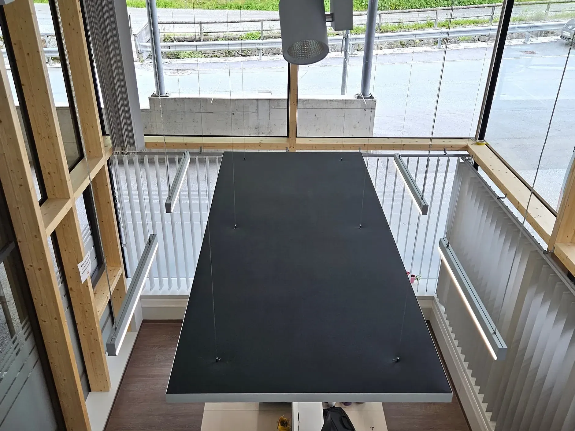 Draufsicht: Großer, schwarzer rechteckiger Tisch in einem Raum mit hellem Holzrahmen. Die Fenster gehen auf eine Straße hinaus.