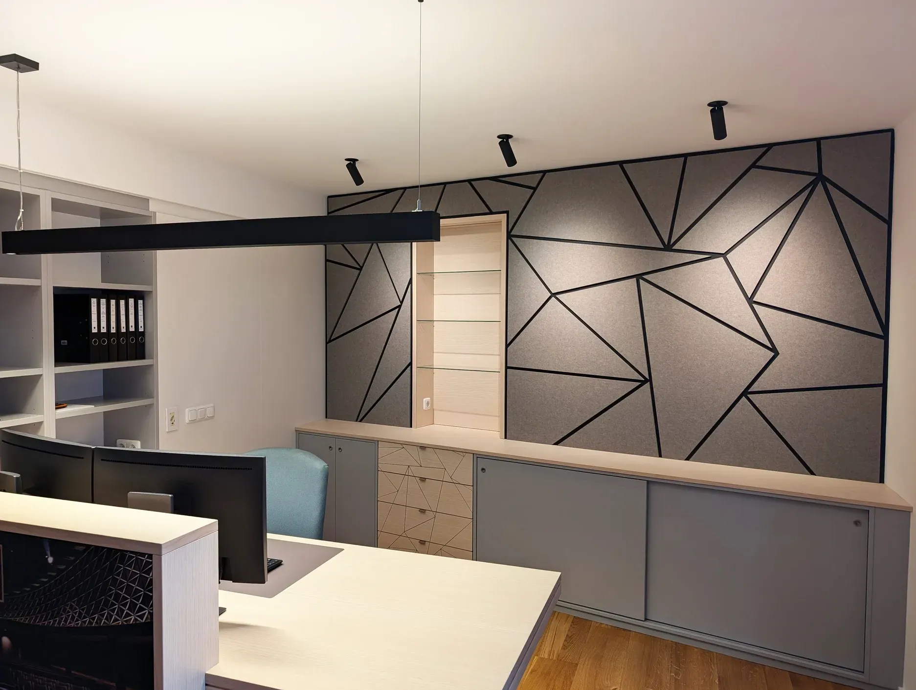 Eine graue, geometrische Akustikwand mit schwarzen Linien umgibt einen hellen, holzvertäfelten Regalbereich in einem Büro