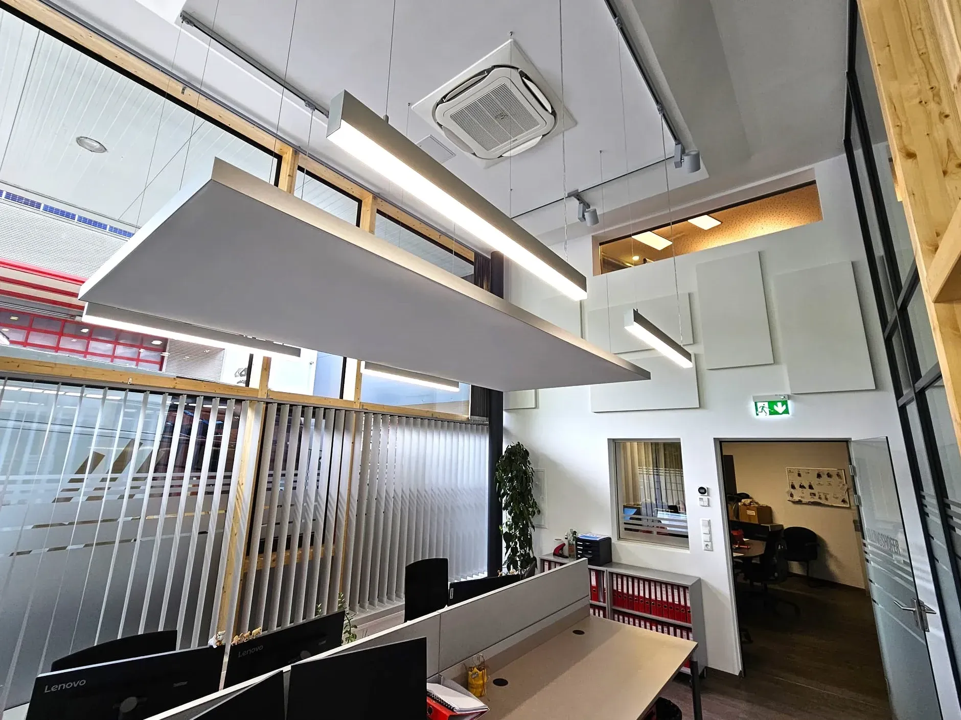 Büroeinrichtung mit Schreibtischen, Computermonitoren und Akustikpaneelen. Deckenbeleuchtung und Fenster.