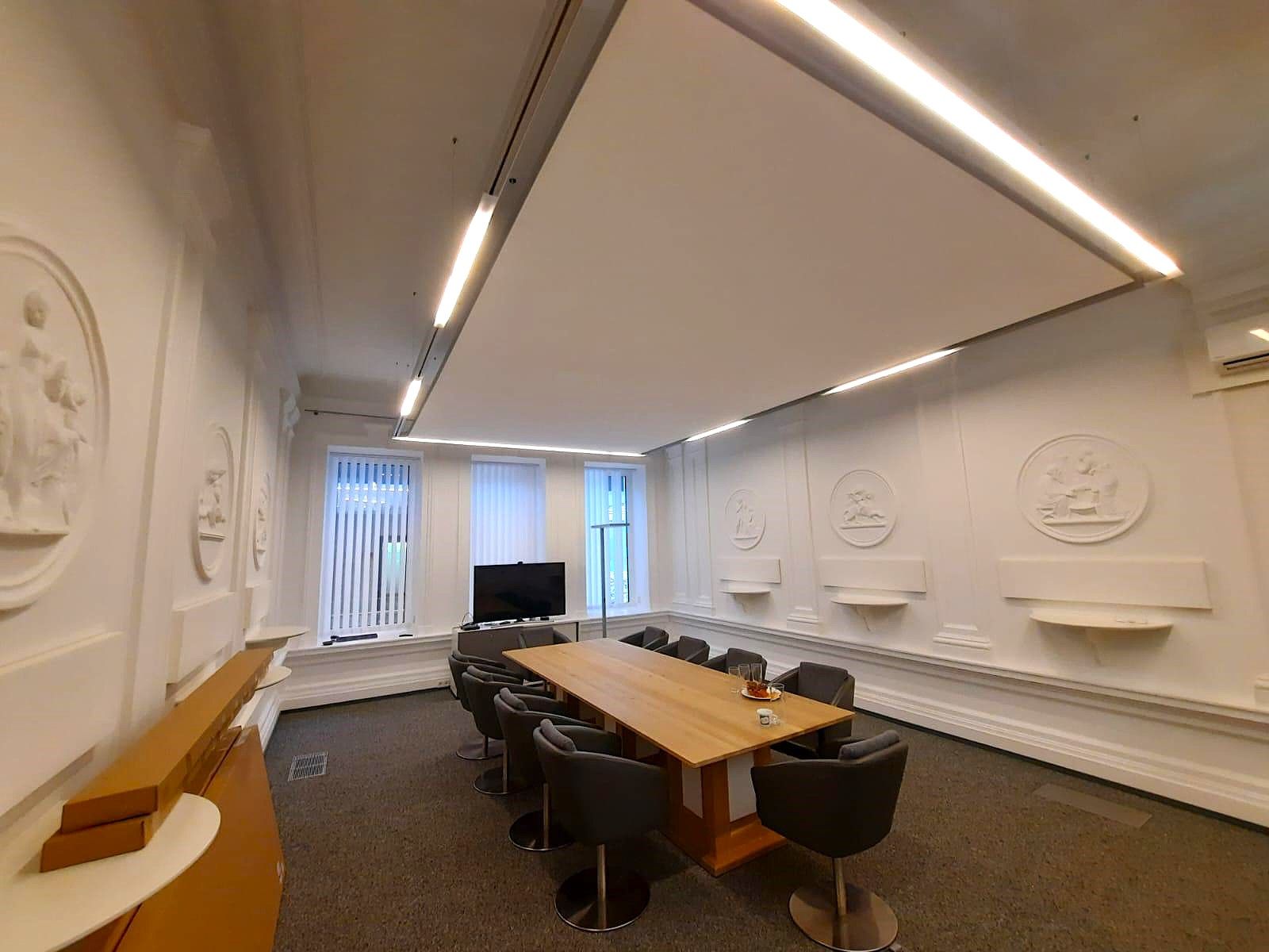 Konferenzraum mit langem Holztisch, schwarzen Stühlen, weißen Akustik Wänden und Zierleisten.