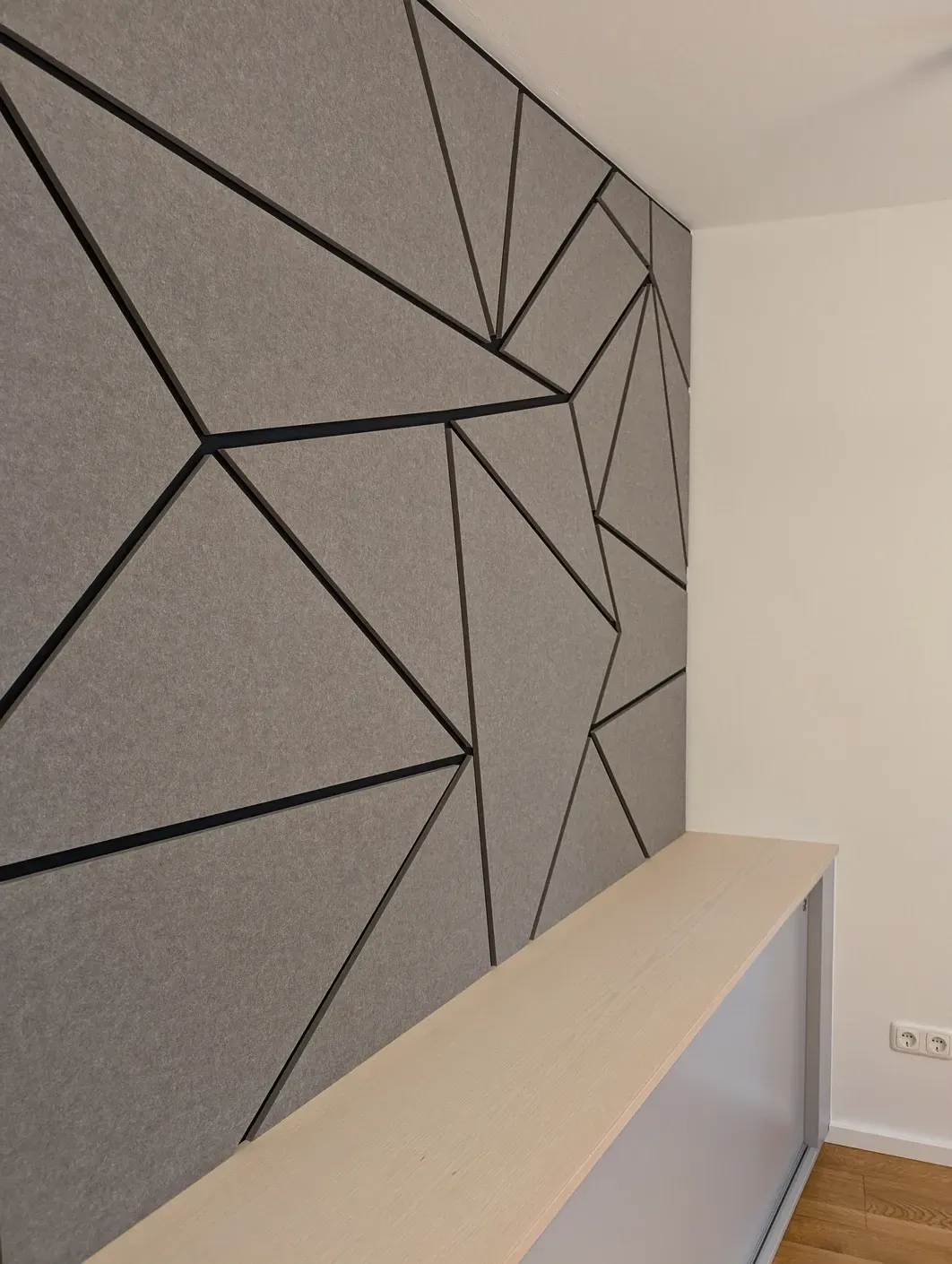 Eine graue, geometrische Akustikwand mit schwarzen Linien umgibt einen hellen, holzvertäfelten Regalbereich.