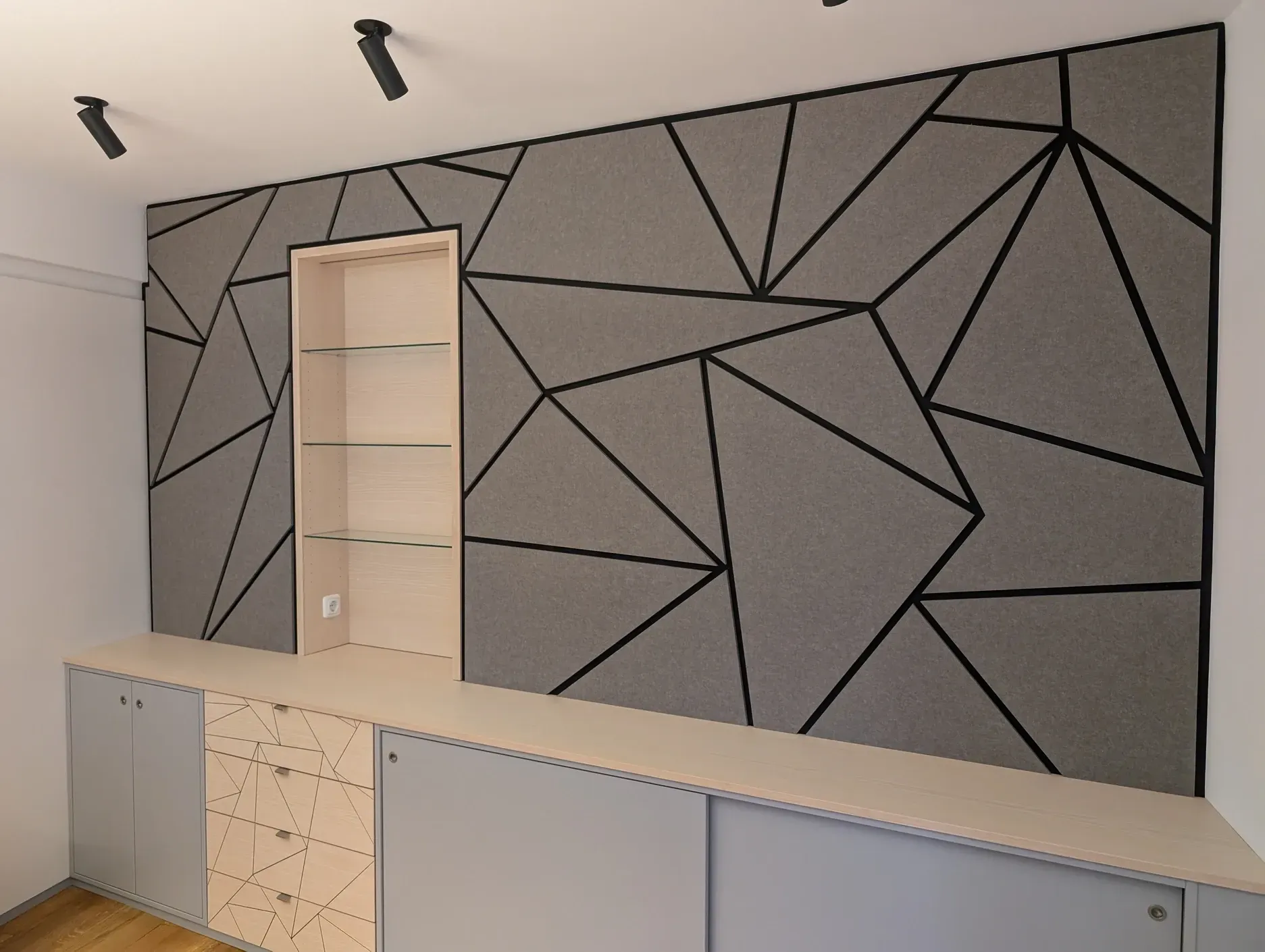 Eine graue, geometrische Akustikwand mit schwarzen Linien umgibt einen hellen, holzvertäfelten Regalbereich.