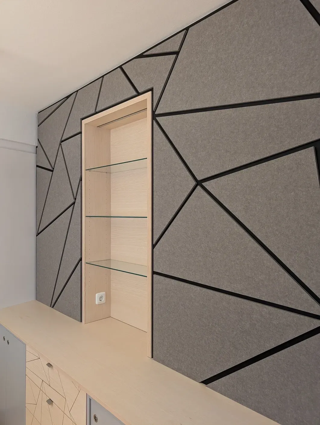 Eine graue, geometrische Akustikwand mit schwarzen Linien umgibt einen hellen, holzvertäfelten Regalbereich.