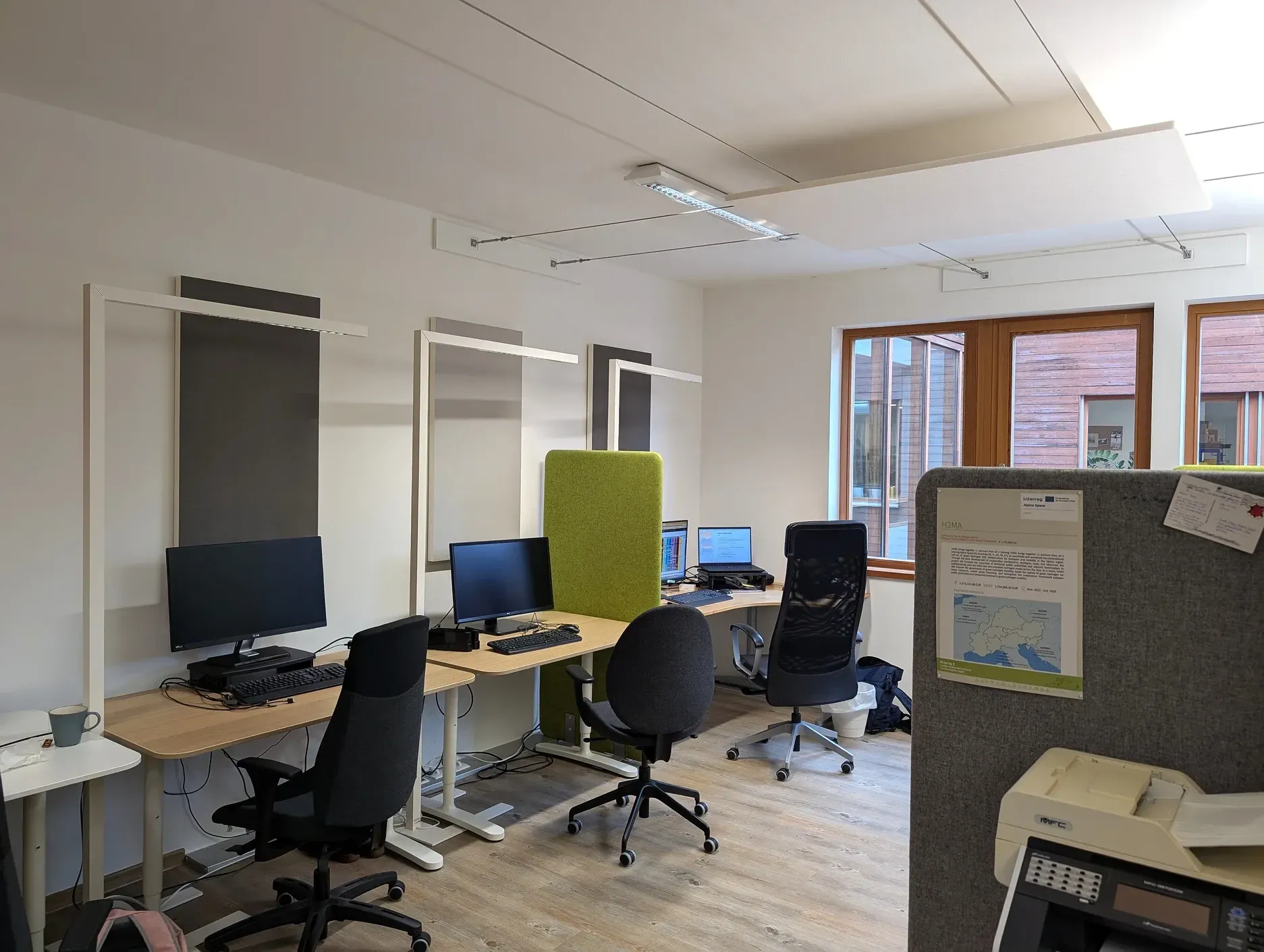 Büro mit Schreibtischen, Computern, Stühlen und Akustikpaneelen; Fenster auf der rechten Seite.