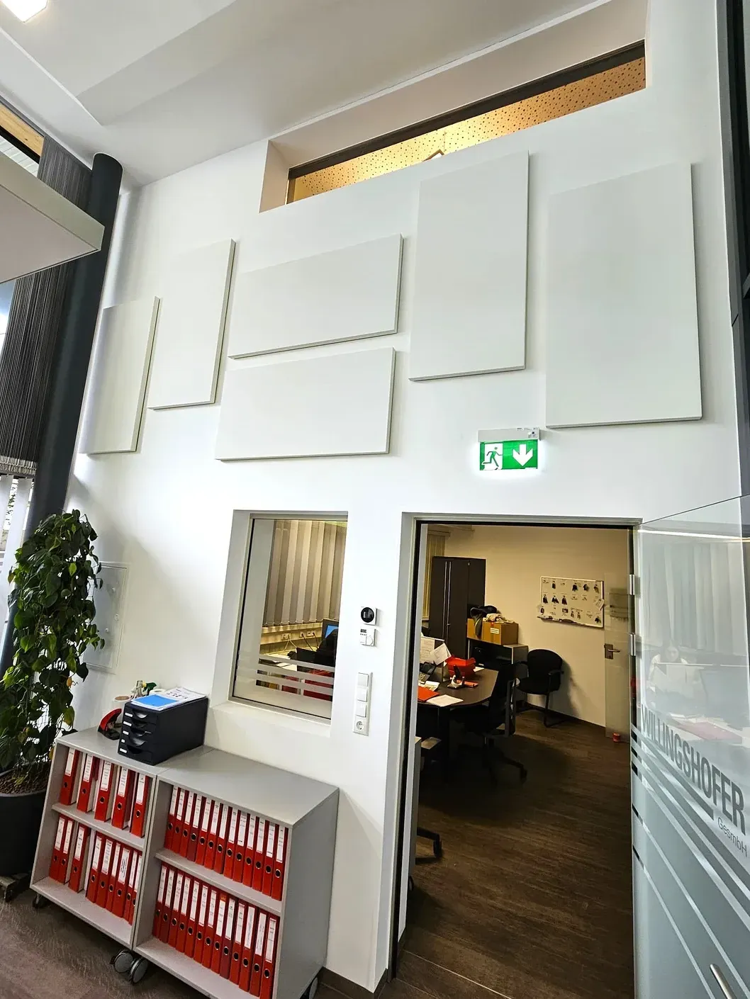 Weiße Akustikpaneele an einer weißen Wand. Ein Büroeingang mit Blick auf Schreibtische und ein Ausgangsschild.