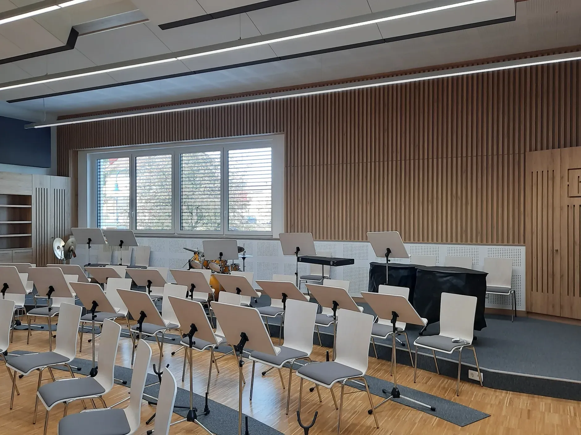 Leerer Musikprobenraum mit Stuhlreihen vor einer kleinen Bühne. Akustik Wänden, Fenster und Deckenleuchten aus Holz.