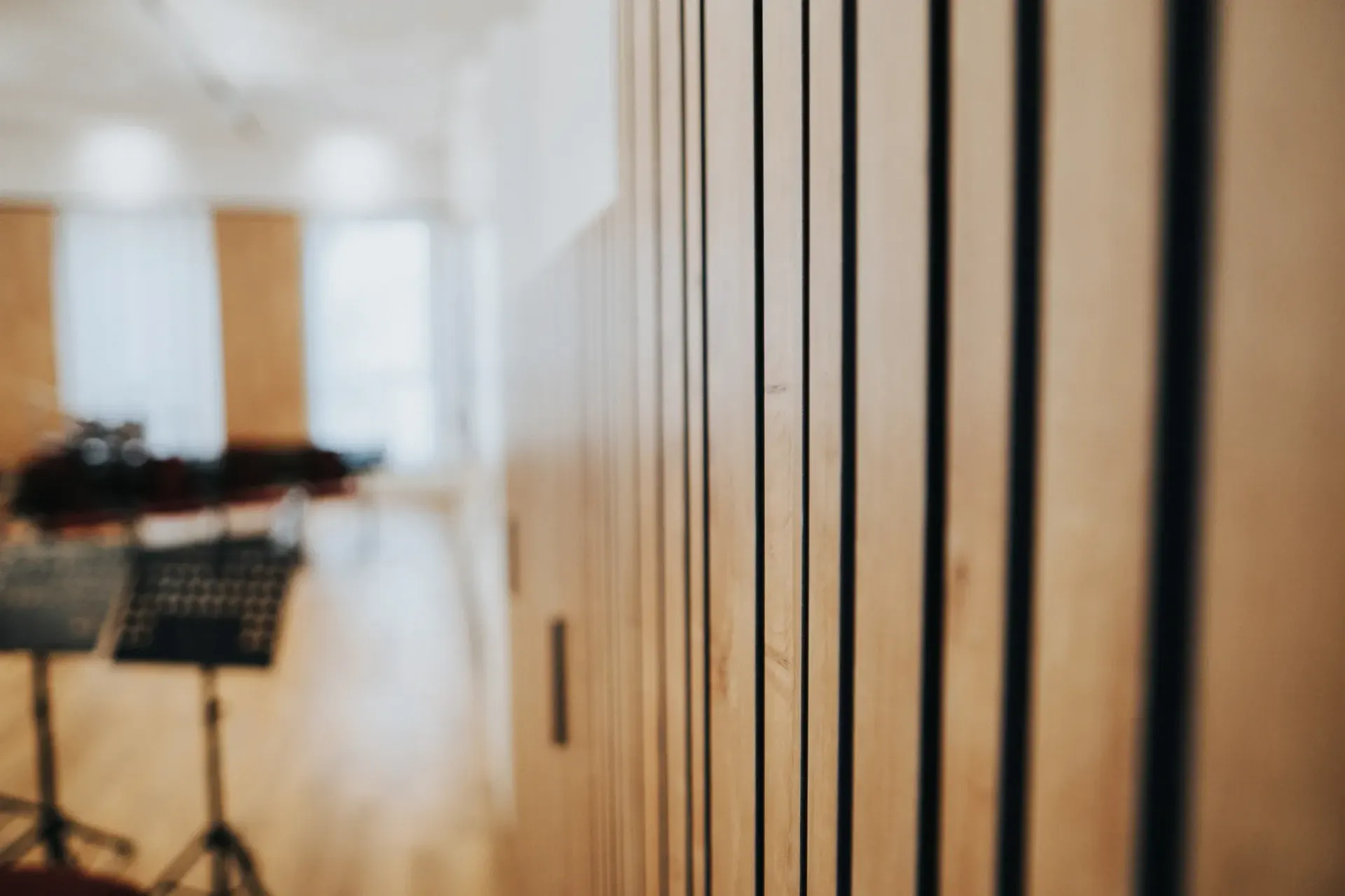 Im Fokus: eine holzgetäfelte Wand mit Akustikpaneelen, im Hintergrund ein verschwommener Musikraum mit Notenständern, natürliches Licht.