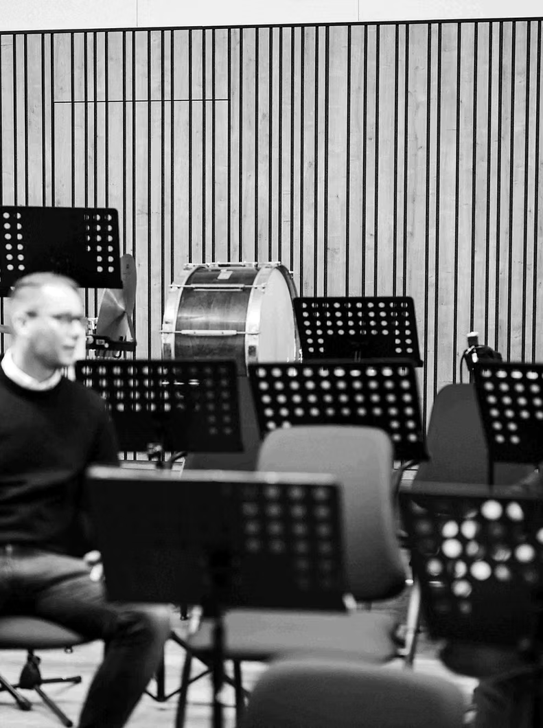 Ein Mann sitzt zwischen Notenständern, im Hintergrund ist eine Basstrommel zu hören, vermutlich in einem Orchesterprobenraum.