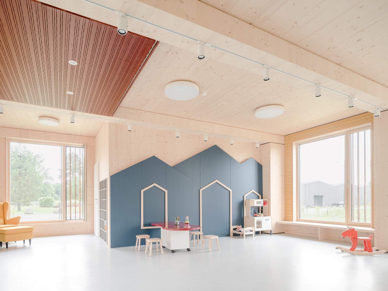 Ein Spielzimmer mit Holzvertäfelung und Akustikpaneelen, großen Fenstern und Spielgeräten vor einer blauen Wand.
