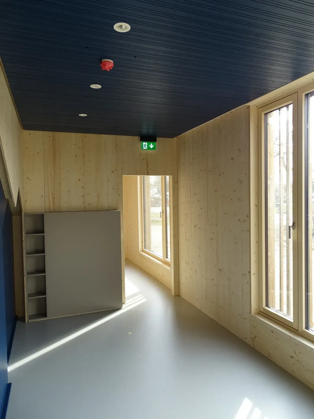 Flur mit blauer Akustik Decke, Holzwänden, grauem Boden und Fenstern; eine Tür führt in einen anderen Raum.