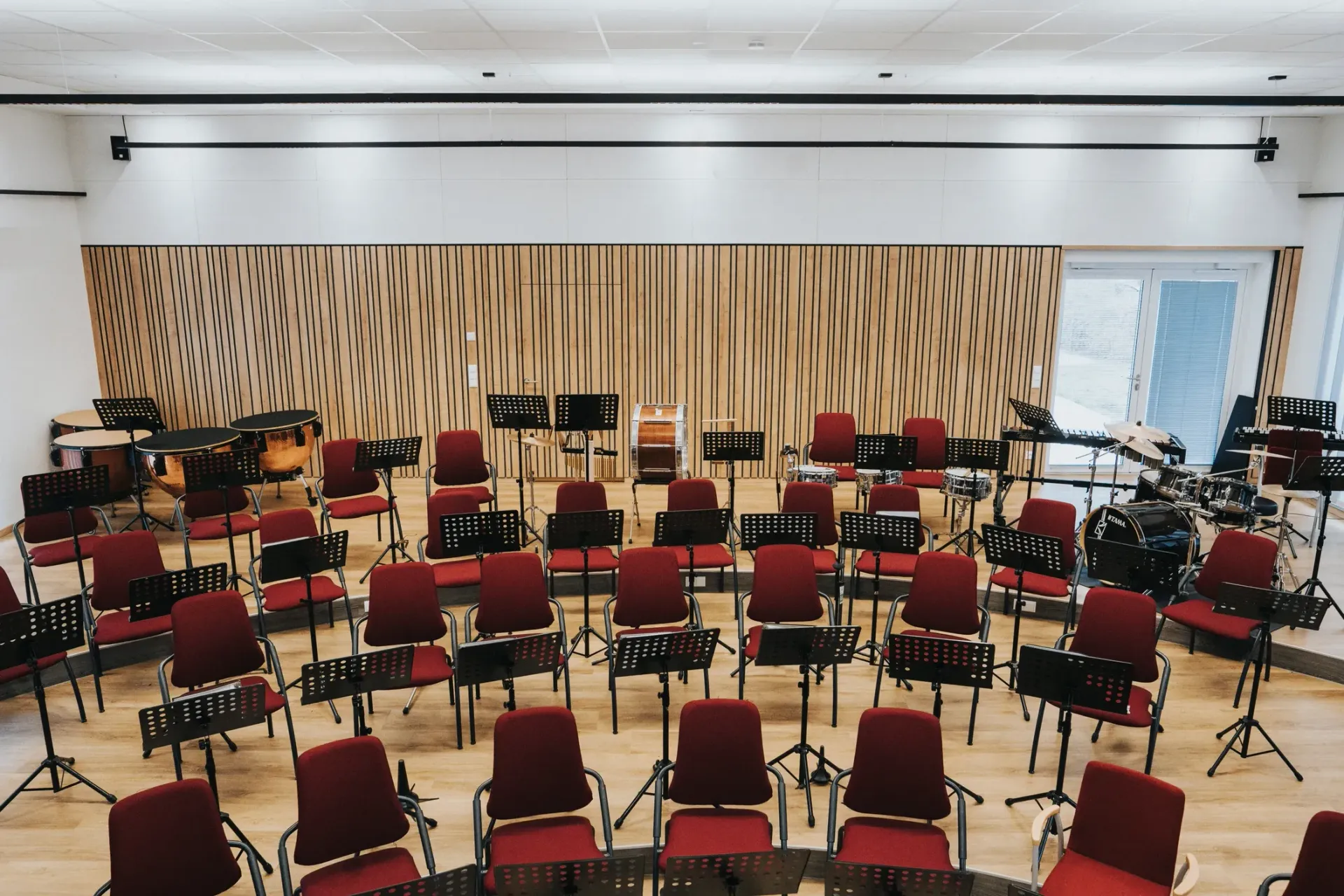 Leerer Orchesterprobenraum mit Akustikpaneelen mit roten Stühlen und Notenständern, die in einem Halbkreis angeordnet sind.
