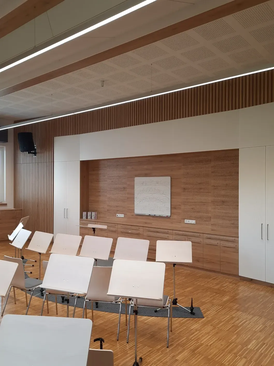 Leerer Konzertsaal mit in Reihen aufgestellten Notenständern, Holzdecke, weißen Wänden und einer Akustikpaneele aus Holz.