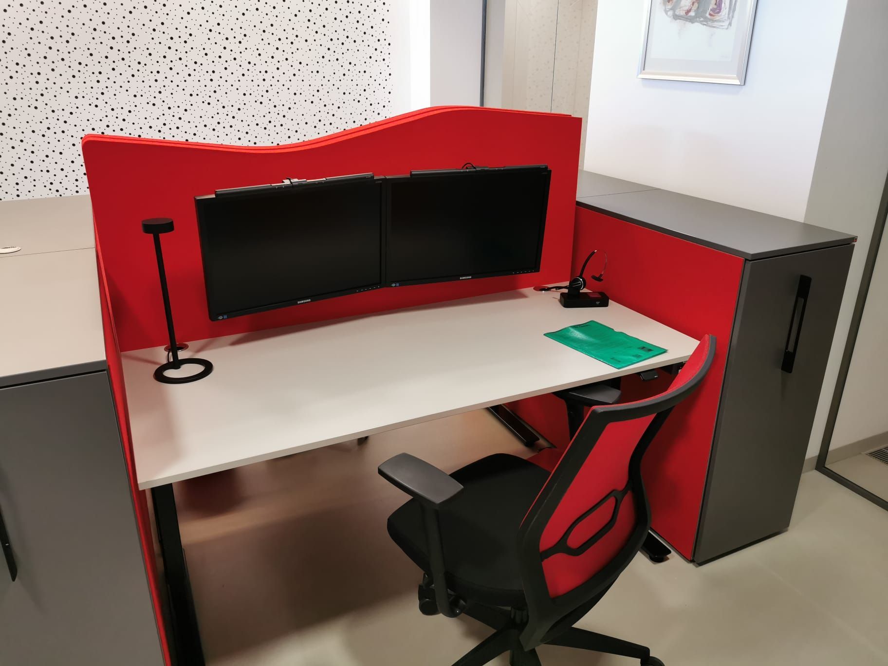 Büroarbeitsplatz mit roter Trennwand, zwei Monitoren, Schreibtisch und schwarz-rotem Stuhl.