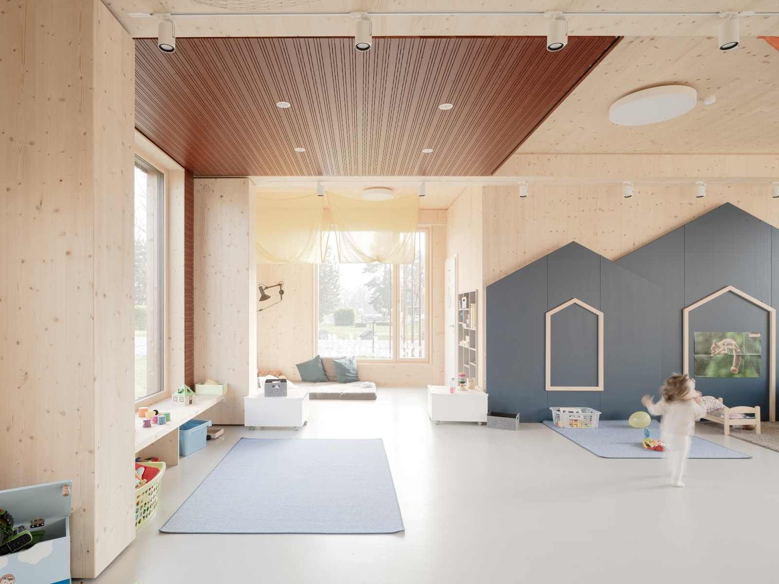 Ein Kind spielt in einem lichtdurchfluteten Zimmer mit Holzwänden und gemusterter Decke; blaue Spielmatten und eine hausförmige Akzentwand.