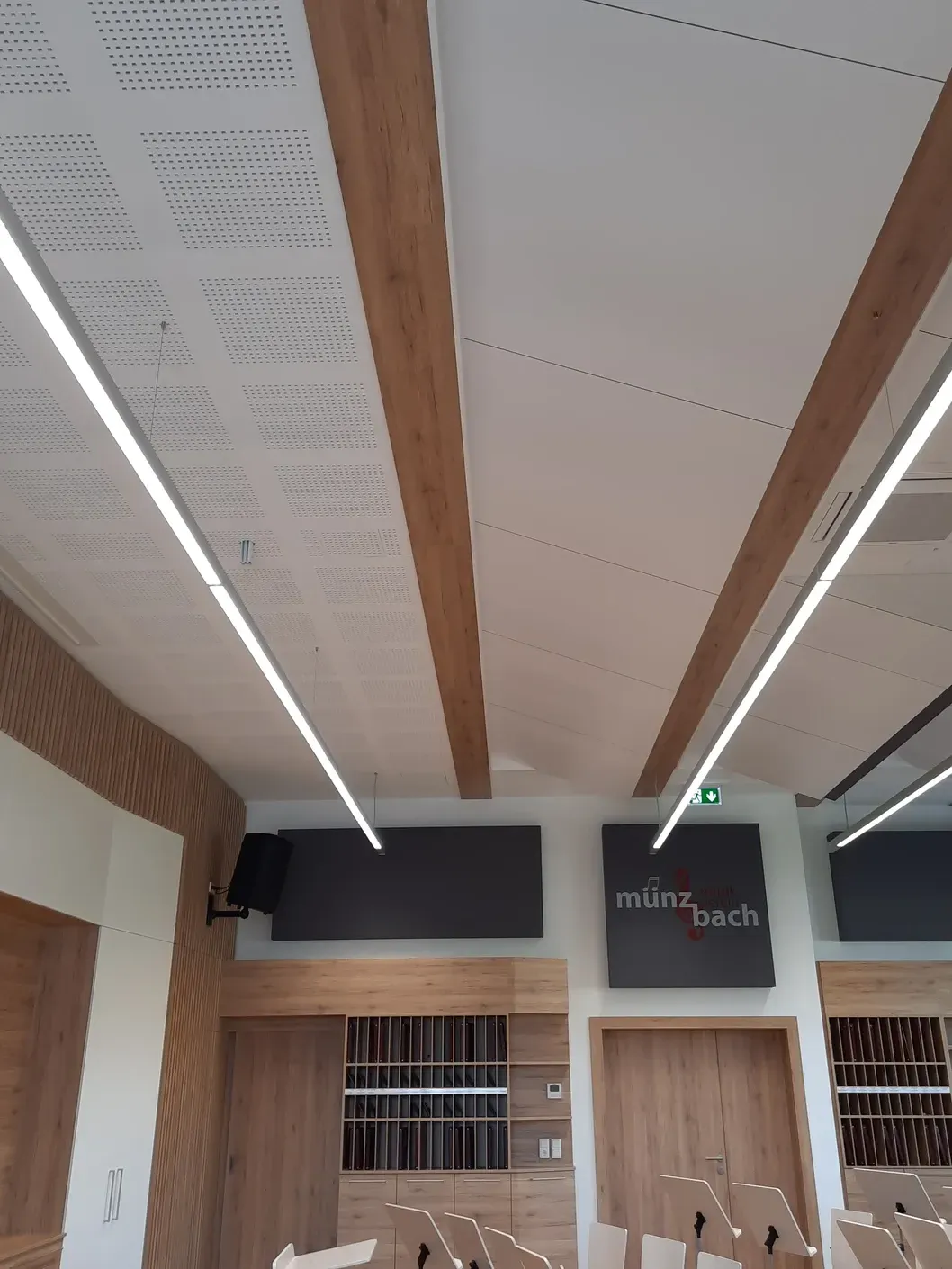 Innenraumdecke mit weißen Paneelen, Holzbalken und linearen Leuchten; ein Besprechungsraum.