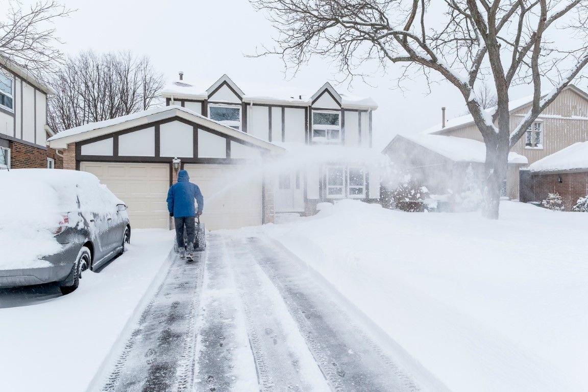 Person in blue jacket shoveling a snowy driveway in front of a house, with parked cars and trees covered in snow.