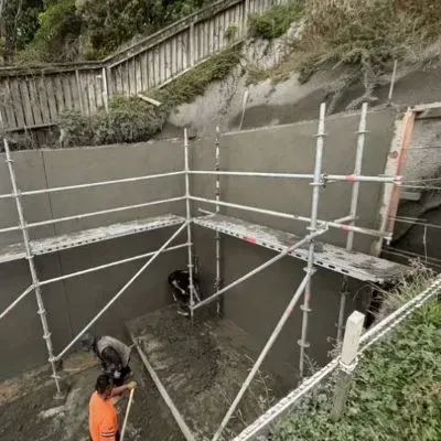 Close-up angle of smooth shotcreted wall inside scaffold bay, workers finishing base slab; Karori, Wellington City.