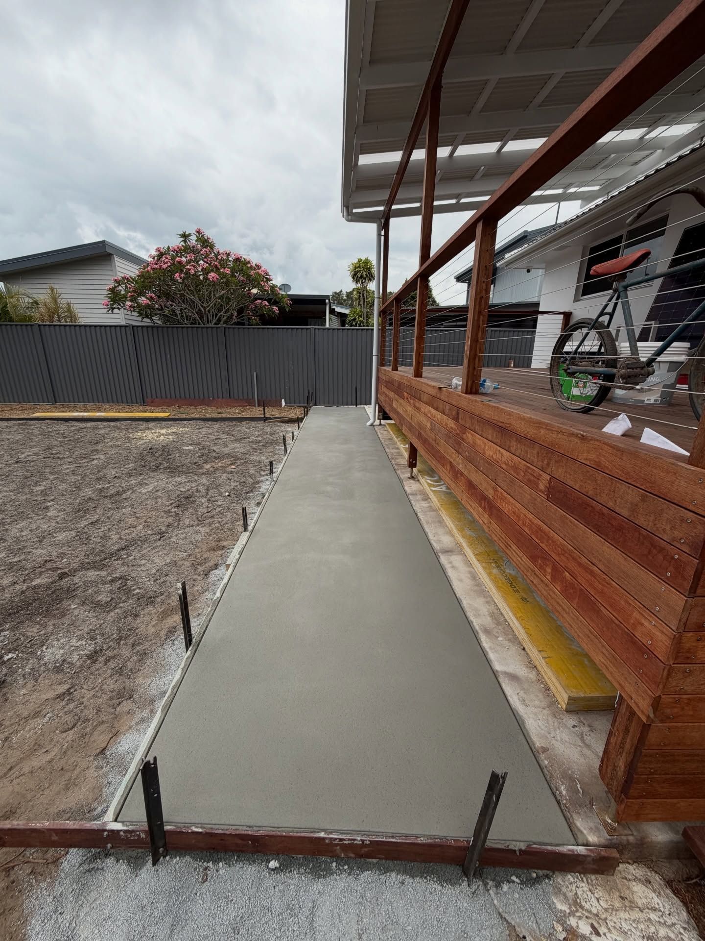 Newly poured concrete walkway next to a wooden deck, bordered by stakes, with a fence and house in the background — All Concrete Creations in Kingscliff, NSW