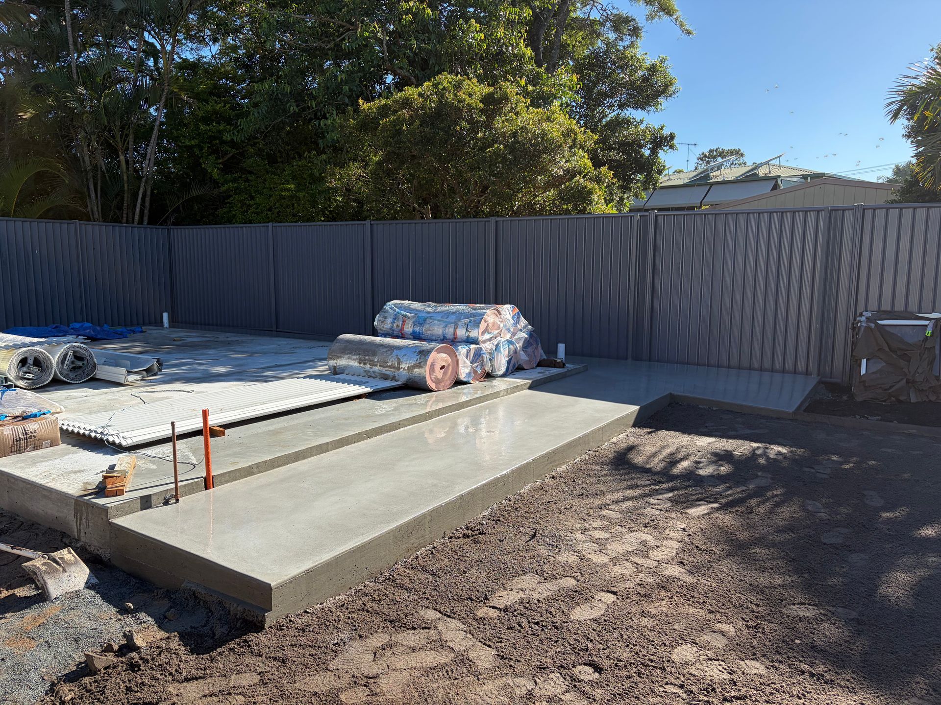 Concrete slab construction site with materials, fence backdrop, and surrounding dirt.