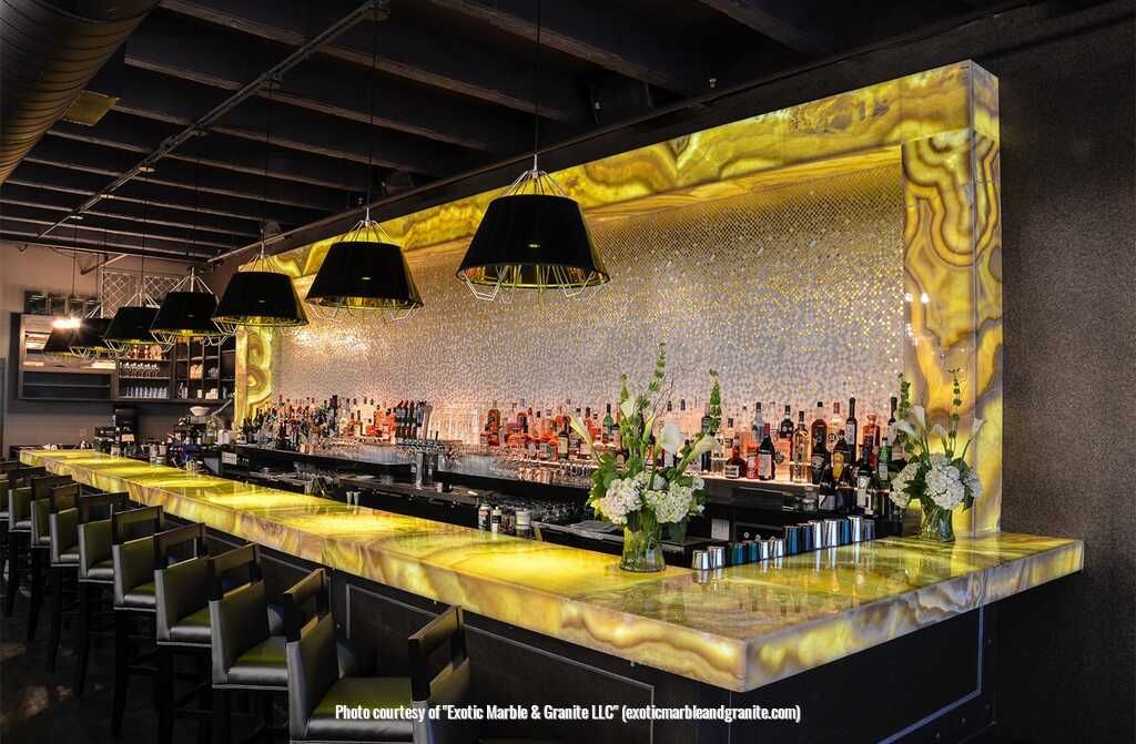 Bar with backlit golden countertop, liquor bottles, black lamps, and seating.