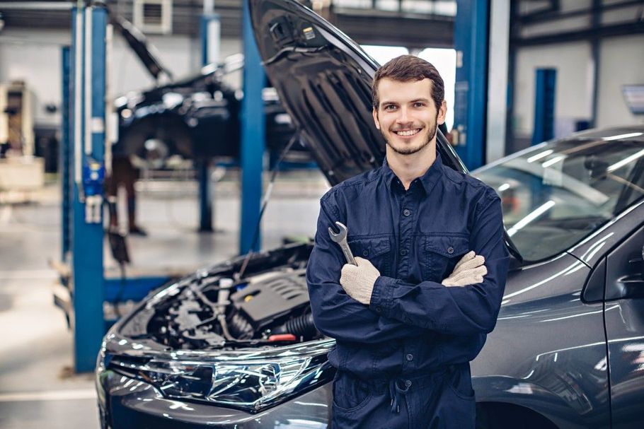 Mechanic in blue coveralls smiling beside a car with its hood open in an auto repair shop