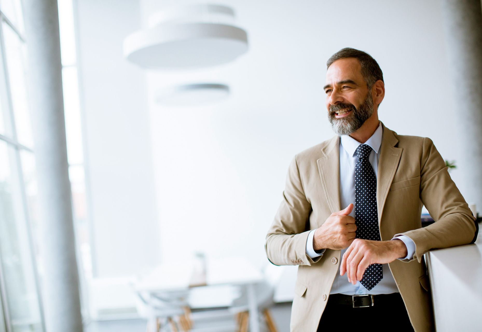 Man in beige blazer and tie, leaning on a white counter, smiling in an office.