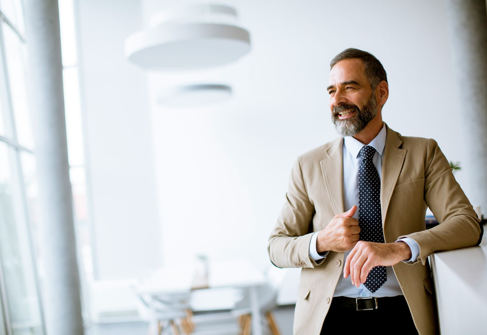 Man in beige blazer and tie, leaning on a white counter, smiling in an office.