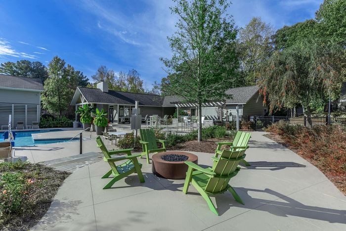 Fire pit with green chairs on a concrete patio, near pool, and clubhouse on a sunny day.