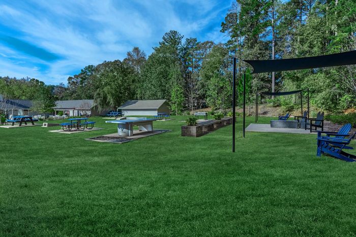 Lush green lawn with outdoor amenities: picnic tables, fire pit, seating, and shade structures near buildings and trees.