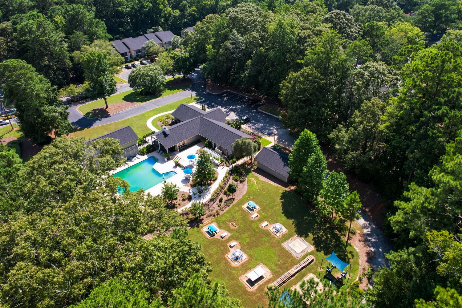 Aerial view of a community pool, buildings, playground, and surrounding trees on a sunny day.