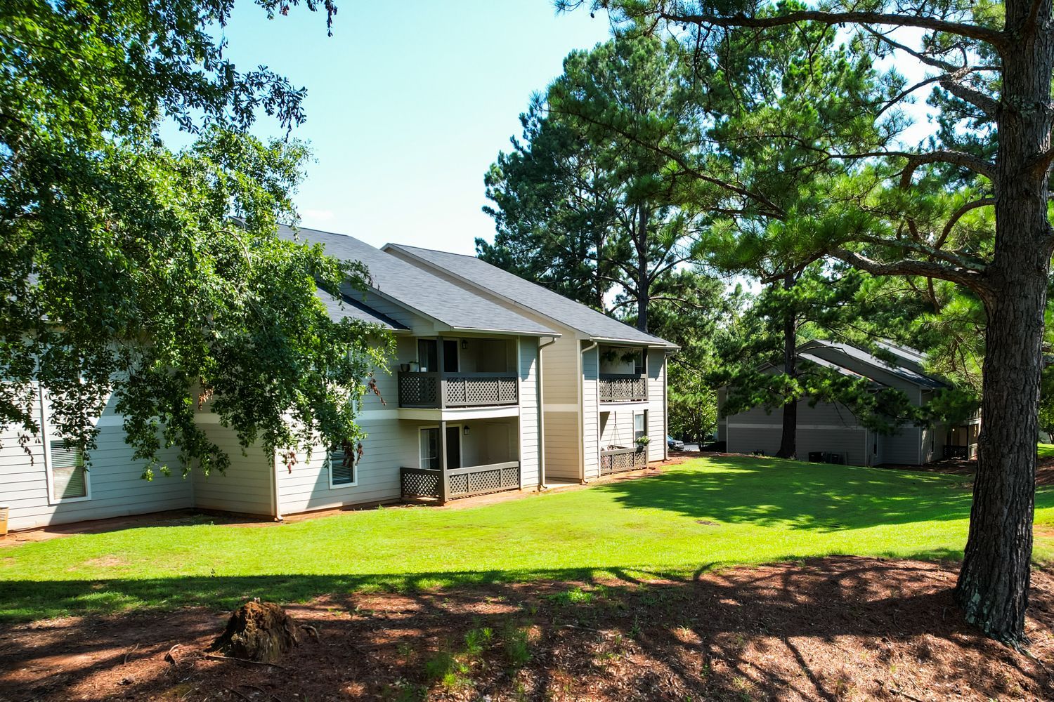 Two-story apartment building with balconies, surrounded by green grass and trees, on a sunny day.