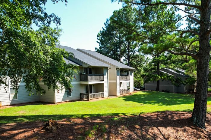 Two-story apartment building with balconies, surrounded by green grass and trees, on a sunny day.
