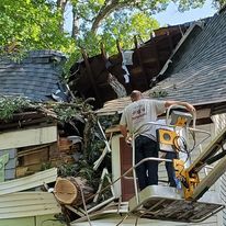 A man is working on the roof of a house that has been damaged by a tree.