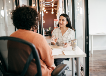 Two people talking at a café table, one facing away and one smiling while gesturing with a glass of water
