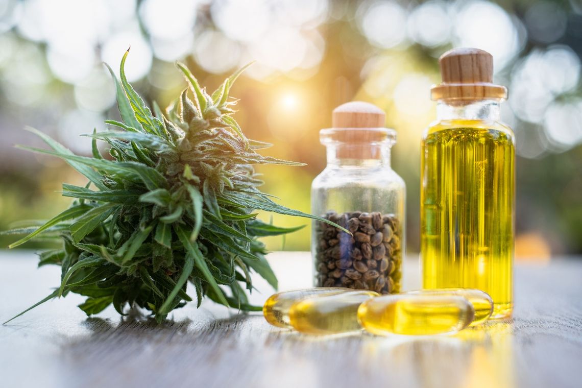 Cannabis bud, oil bottles, capsules, and peppercorns on a wooden surface with a blurred outdoor background