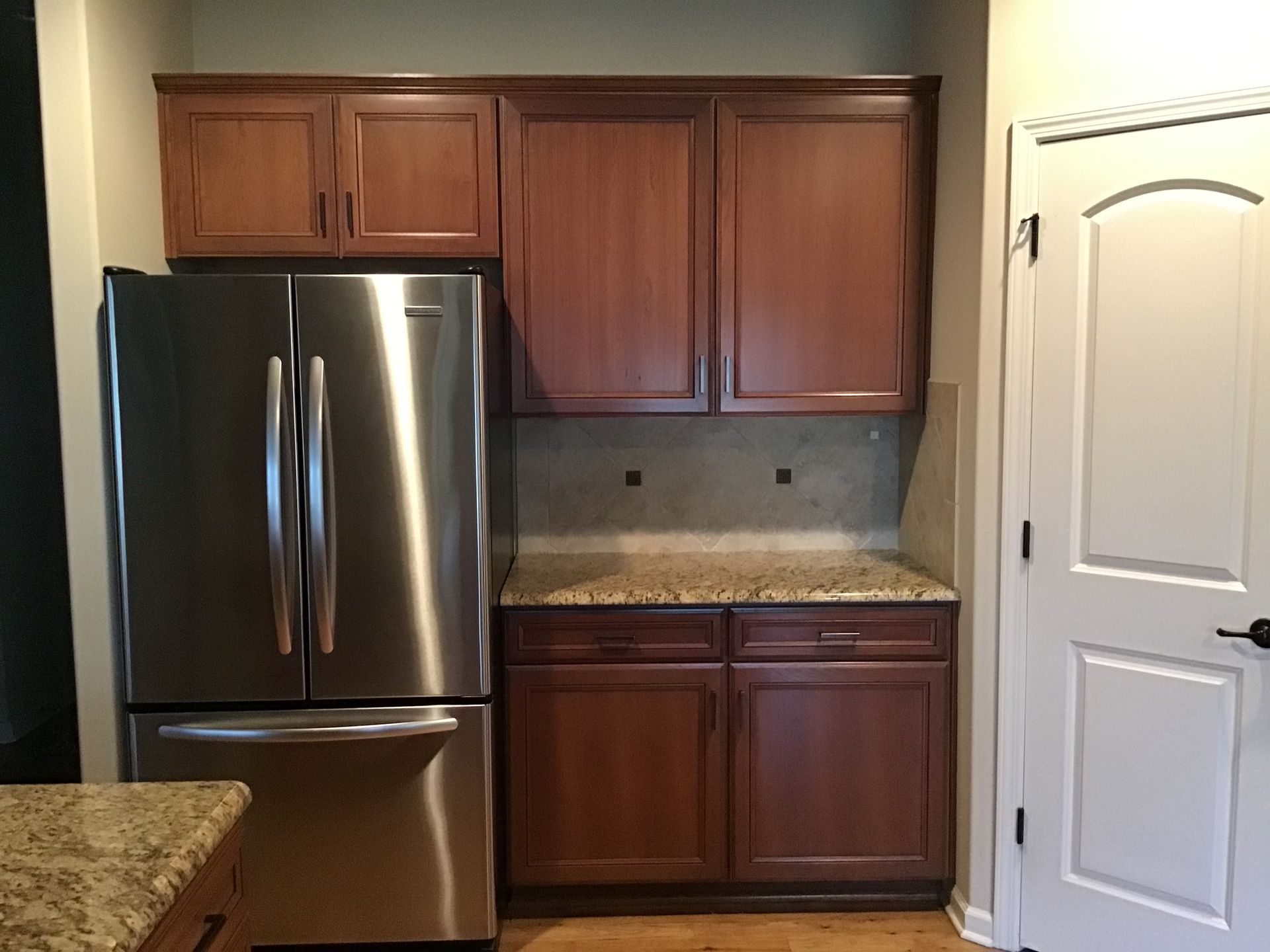 Small kitchen with stainless steel fridge, wood cabinets, granite counters, and a white door.