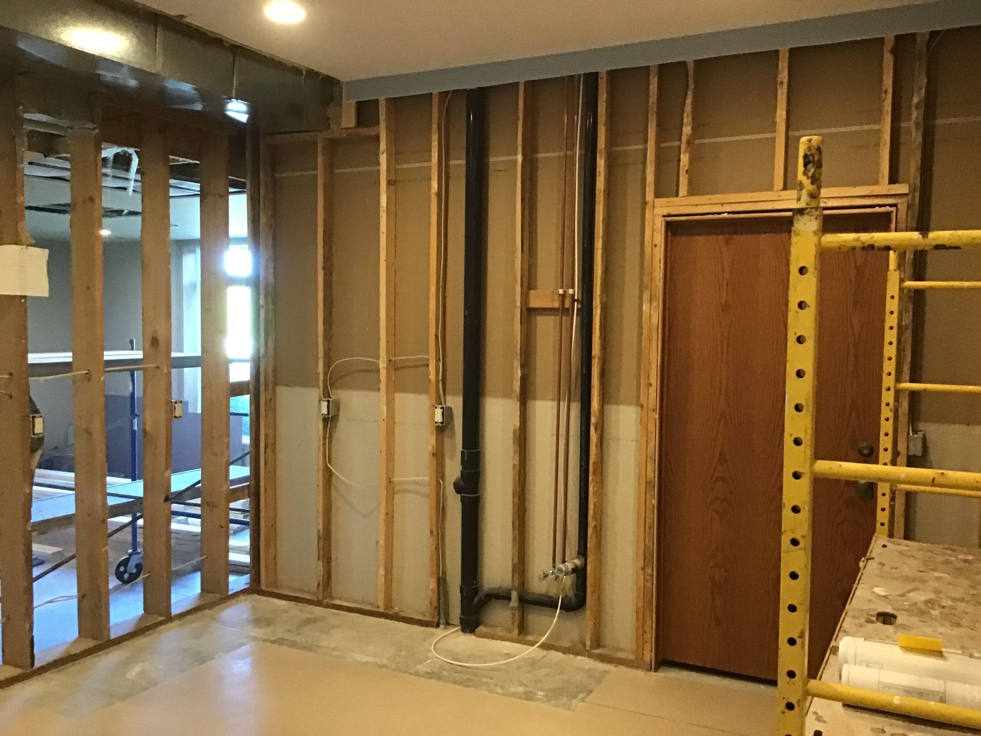 Bare interior room under renovation with exposed studs, pipes, and a yellow scaffold on the right.