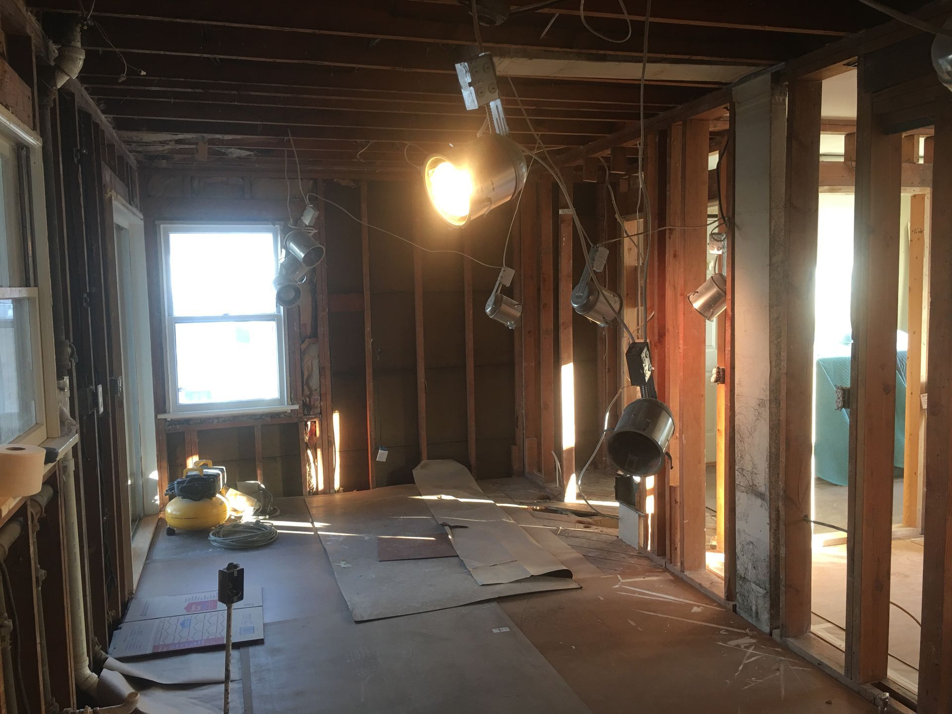 Damaged, unfinished room with exposed studs, broken floorboards, debris, and a single lit bulb near a window