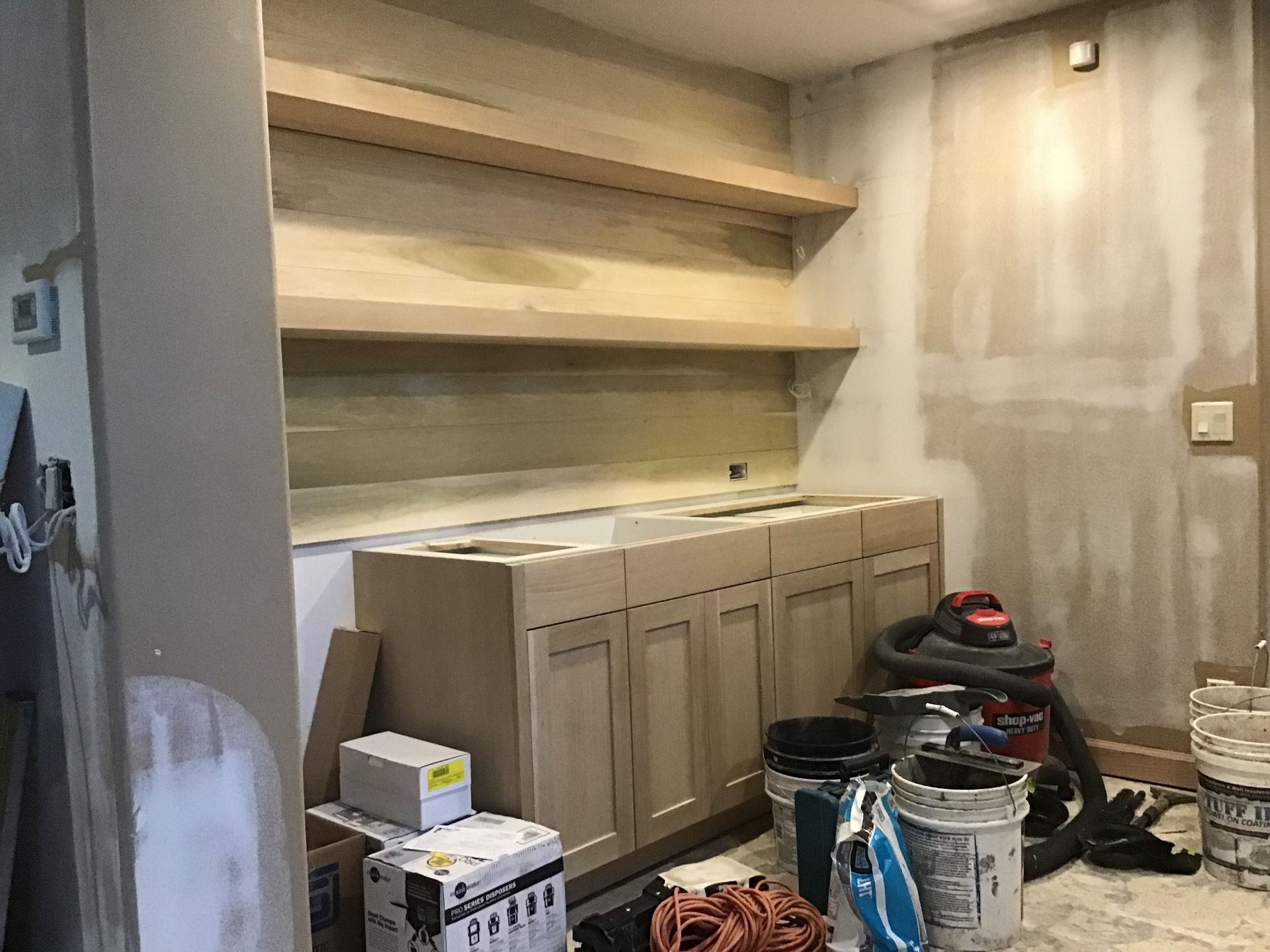 Unfinished kitchen alcove with cabinets, open shelves, and painting supplies on the floor.