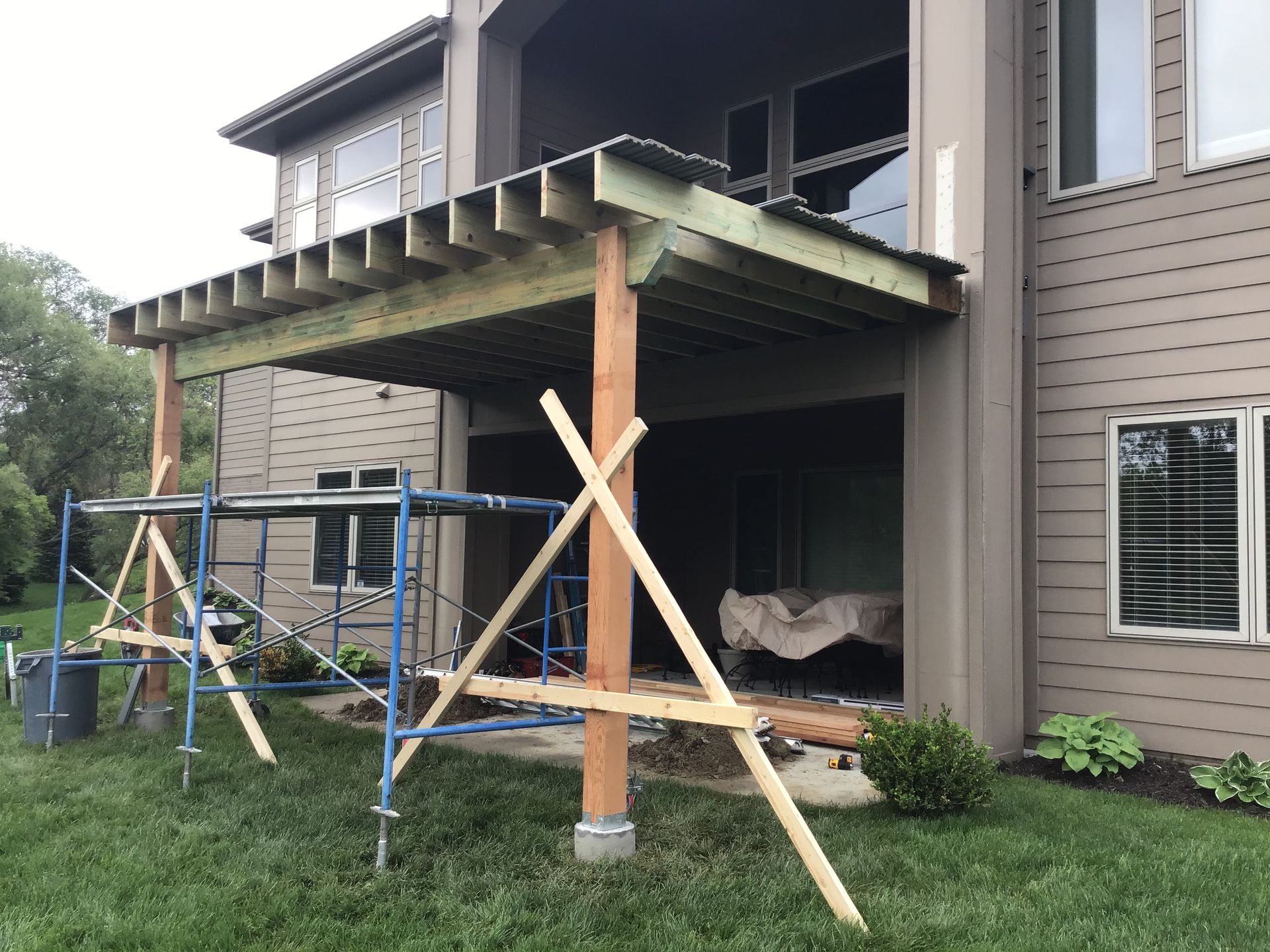 Partially built porch roof with wood beams and blue scaffolding beside a beige house