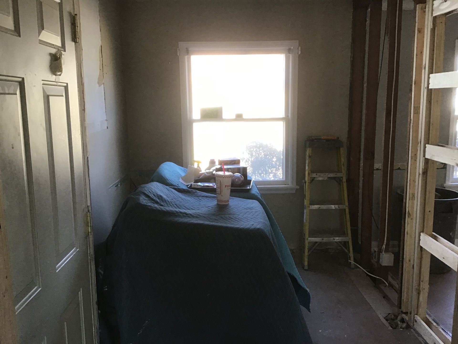 Dim room with a bed beneath a bright window and unfinished walls, plus a ladder on the right.