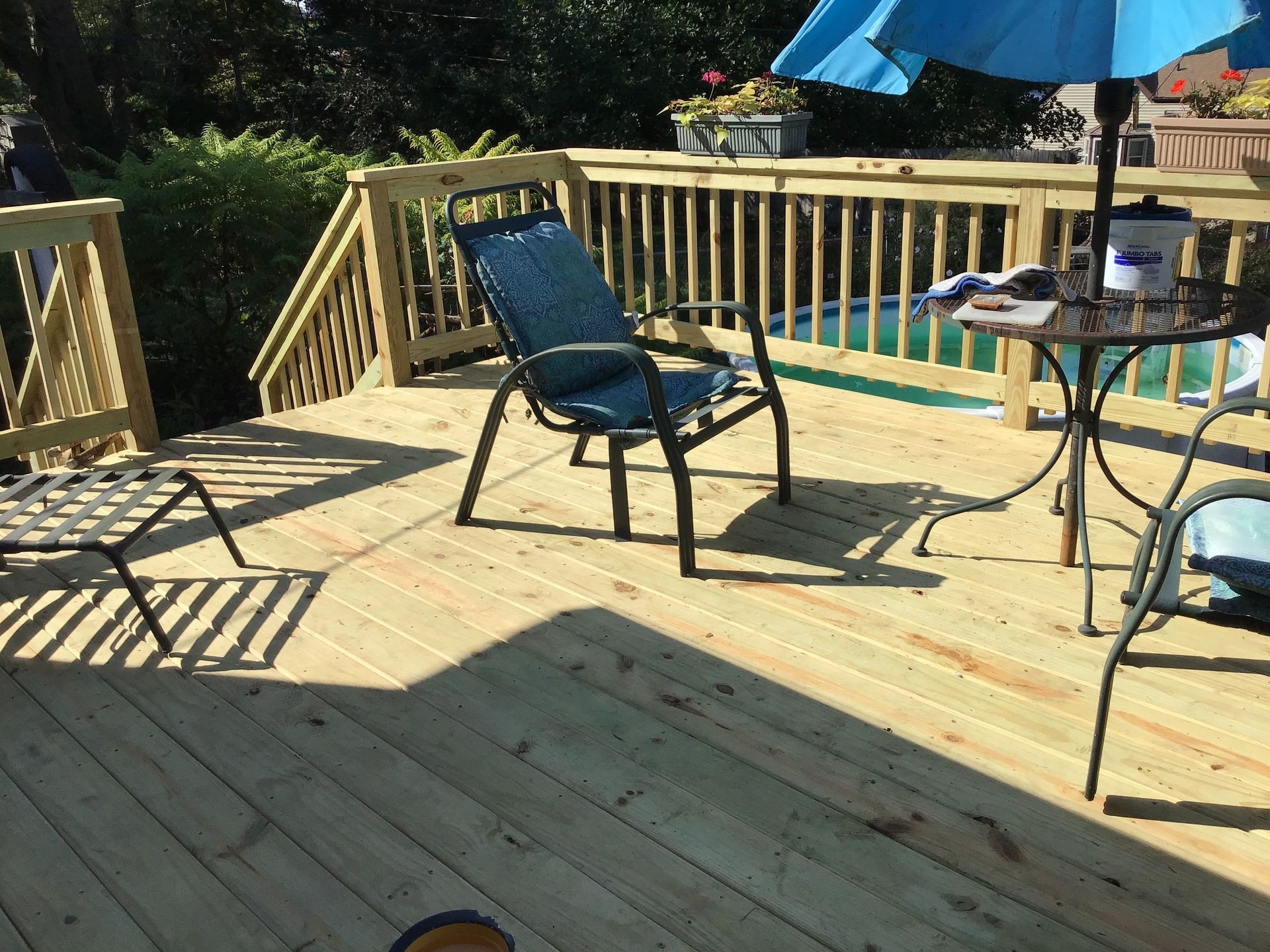 Wooden deck with black patio chairs, small table, and blue umbrella in a sunny backyard.