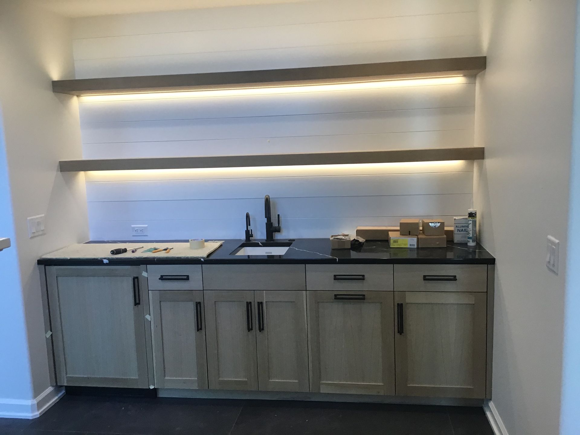 Kitchen bar area with gray cabinets, black countertop, and wall-mounted shelves under warm lighting