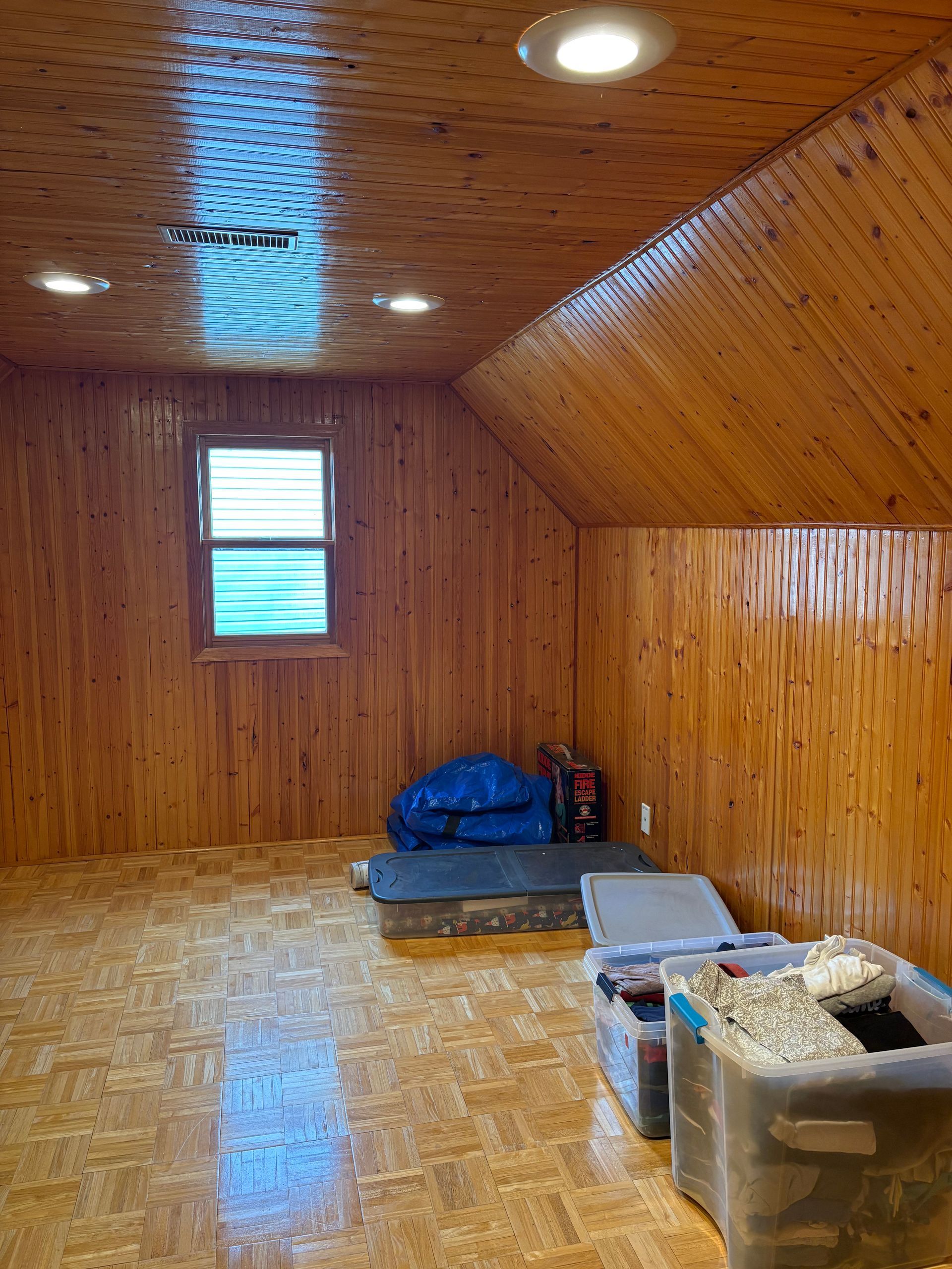 Small wood-paneled room with a sloped ceiling, window, blue bags, and storage bins on the floor.