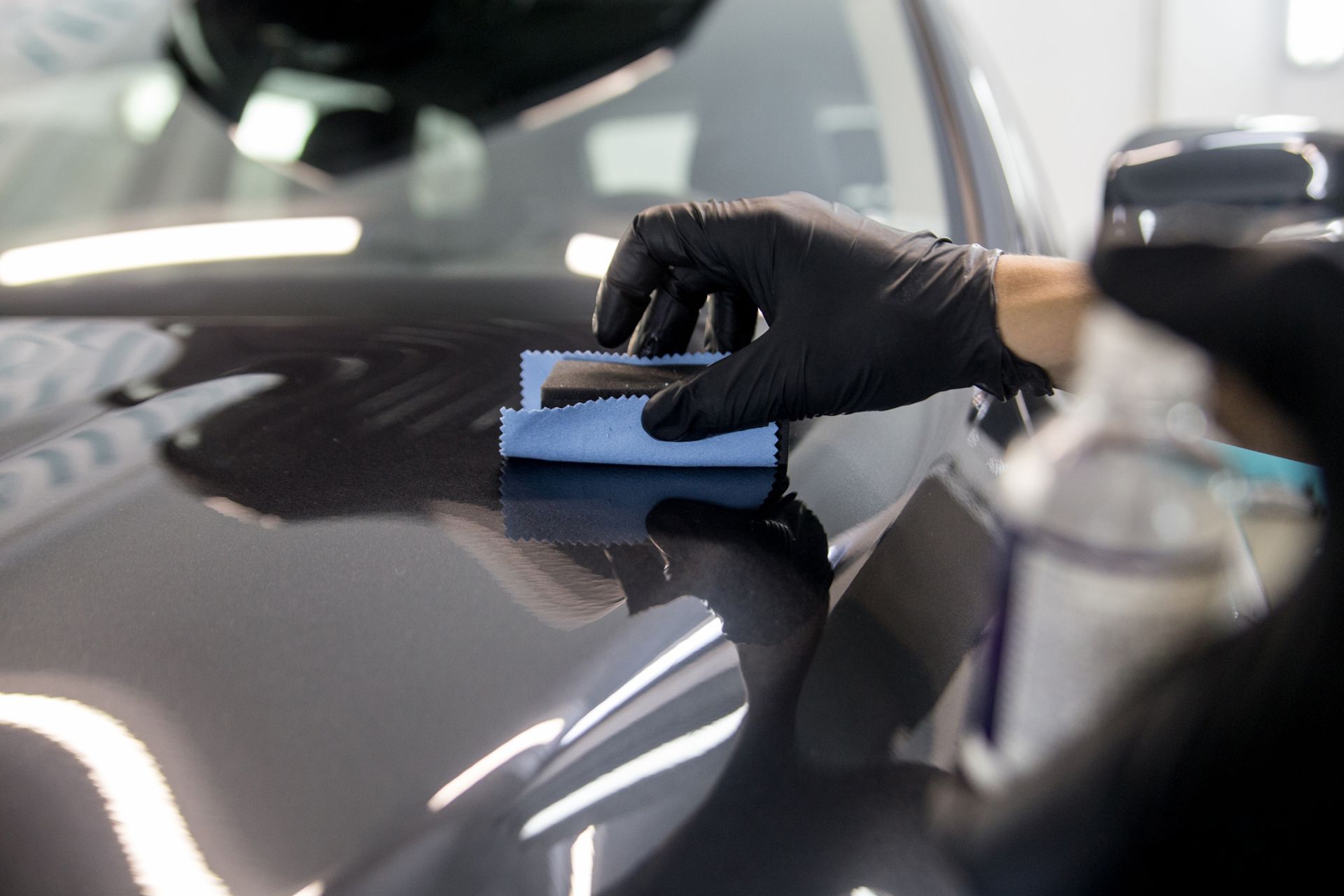 A gloved hand uses a blue applicator sponge to apply ceramic coating to the glossy hood of a dark car.
