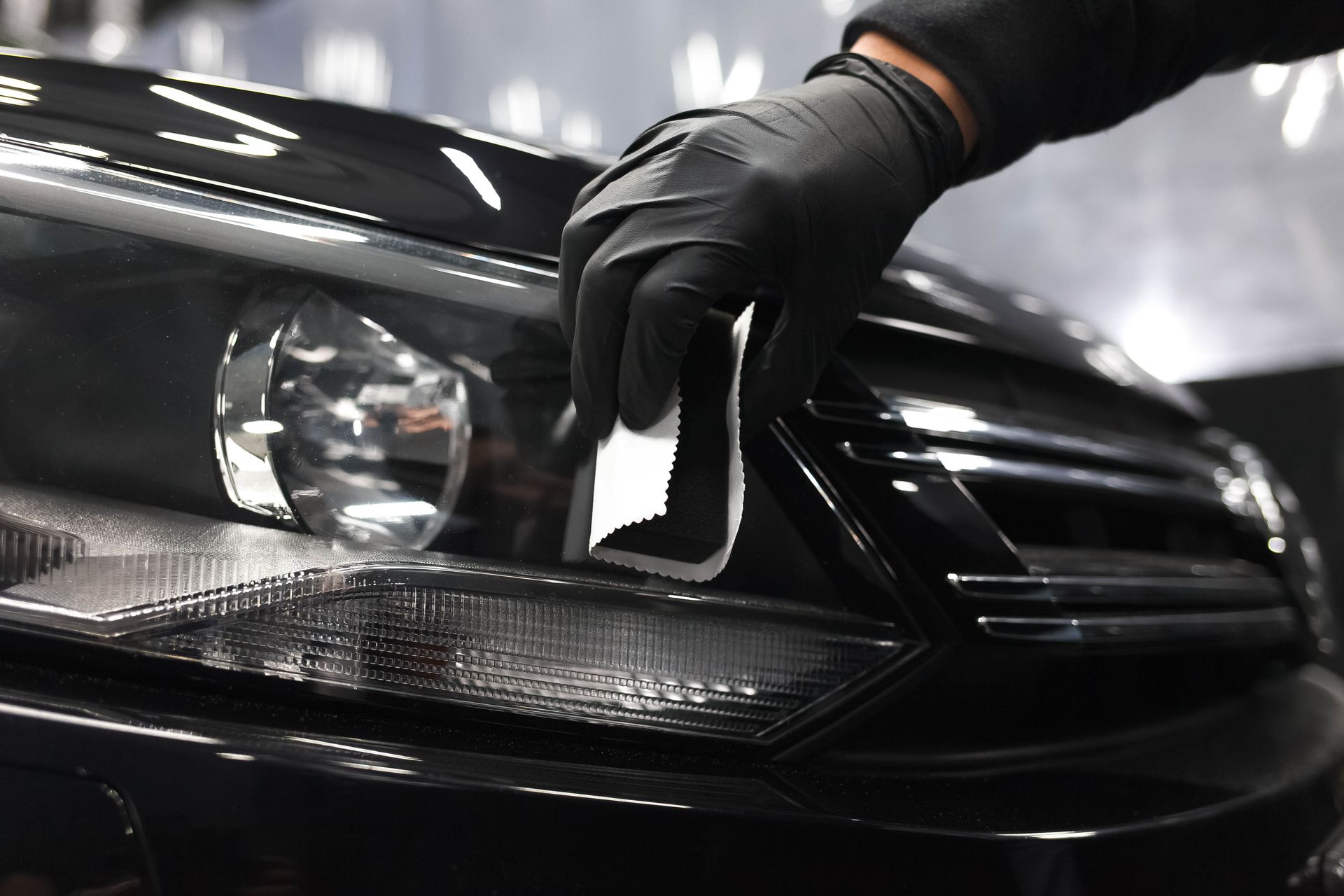 A person in a black glove uses a cloth to clean a car's black headlight.