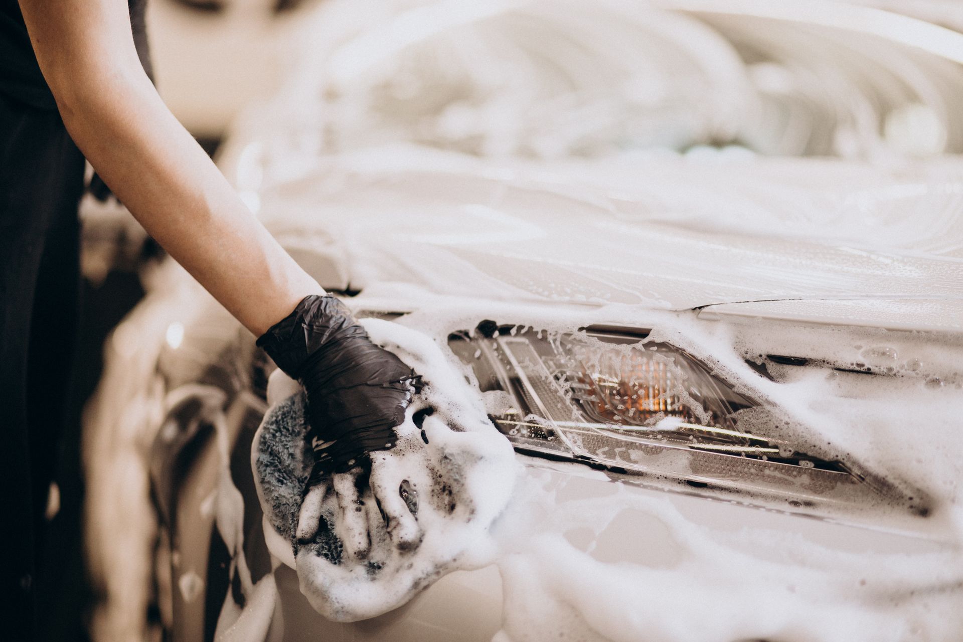 A person wearing a black glove uses a soapy sponge to clean the headlight of a car.